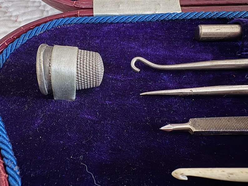 May include: A vintage sewing kit with a silver thimble, needles, and crochet hooks, set against a deep purple velvet background. The tools are neatly arranged within a red and blue-trimmed case, showcasing craftsmanship.
