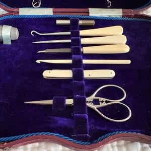 May include: An antique sewing kit with a deep purple velvet interior. The kit contains various tools, including a thimble, hooks, needles, a small knife, and ornate scissors. The case is trimmed with red and blue accents.