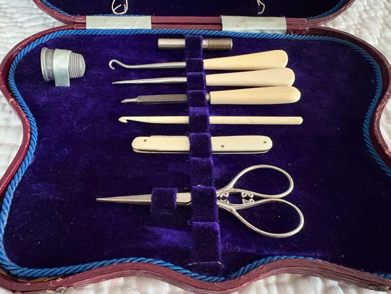 May include: An antique sewing kit with a deep purple velvet interior. The kit contains various tools, including a thimble, hooks, needles, a small knife, and ornate scissors. The case is trimmed with red and blue accents.
