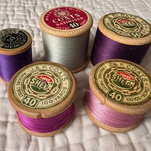 May include: A collection of vintage thread spools in various colors, including purple, pink, and gray. The spools have wooden bases and feature labels with text such as "J. & P. Coats Super Sheen" and "Machine Twist".