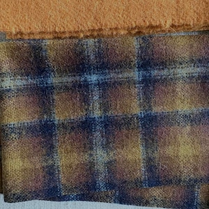 May include: Close-up of fabric swatches. The primary swatch features a plaid pattern in shades of blue, gold, and brown. Above it is a solid orange-brown fabric. The fabrics have a textured appearance, suggesting a woven material.