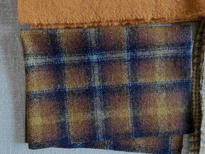 May include: Close-up of fabric swatches. The primary swatch features a plaid pattern in shades of blue, gold, and brown. Above it is a solid orange-brown fabric. The fabrics have a textured appearance, suggesting a woven material.