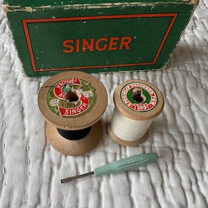 May include: A green and orange cardboard box labelled SINGER, two wooden spools of thread with vintage labels, one with black thread and one with white thread, and a light green metal screwdriver with the word SINGER embossed on the handle.
