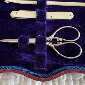 May include: A vintage sewing kit with a thimble, scissors, a crochet hook, and a small knife. The tools are arranged on a deep purple velvet background. The scissors have ornate handles.
