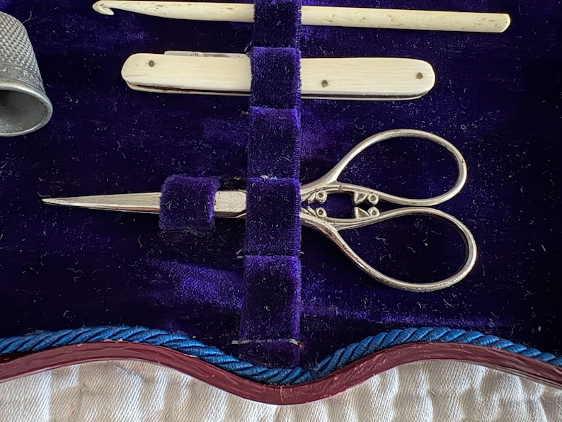 May include: A vintage sewing kit with a thimble, scissors, a crochet hook, and a small knife. The tools are arranged on a deep purple velvet background. The scissors have ornate handles.