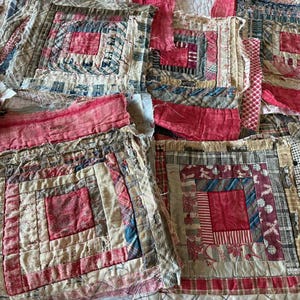 May include: A collection of square fabric quilt blocks featuring a log cabin pattern. The blocks are constructed from various textile scraps in shades of red, cream, blue, and grey, showcasing diverse patterns including stripes, plaids, and floral prints.