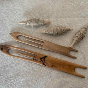 以下が含まれることがあります： 木製の織り道具と糸巻きが、質感のある表面に配置されています。2つの木製のシャトルと、さまざまなサイズの3つの糸巻きが見えます。シャトルは明るい茶色です。