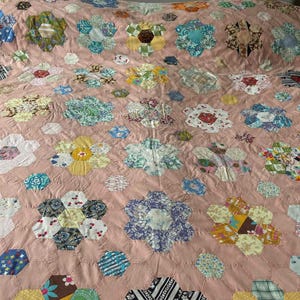 May include: A vintage quilt with a pink base and a floral design. The quilt showcases numerous hexagonal flower shapes in various colours, including blue, yellow, and white. The quilt is laid out on a bed.