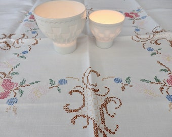 Puede incluir: Dos portavelas de cerámica blanca con un diseño texturizado, colocados sobre un mantel blanco con un patrón de bordado floral.