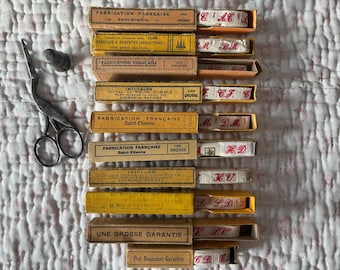 May include: A collection of ten rectangular cardboard boxes containing rolls of white fabric tape with red embroidered initials, alongside a pair of metal stork-shaped embroidery scissors and a small metal thimble for sewing projects.