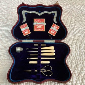May include: An open antique sewing kit with a burgundy and blue exterior. Inside, a deep blue velvet lining holds various sewing tools, including needles, scissors, and ivory-colored handles. The kit includes small boxes of "Sharps" needles and a label that reads "English Make Art Needles."