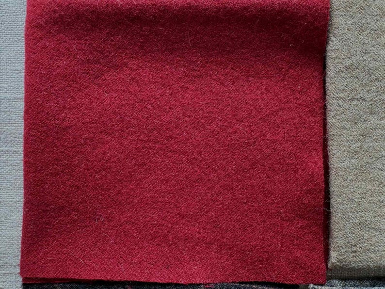 May include: Close-up of three fabric swatches. The primary swatch is a deep red, with a textured surface. Adjacent to it is a gray-green swatch, and a light gray swatch is partially visible on the left. The fabrics appear to be wool or a similar material.