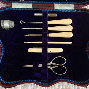 May include: A vintage sewing kit in a burgundy case with a blue velvet interior. The kit includes a thimble, scissors, and various ivory-handled tools. The case is trimmed with blue cording.