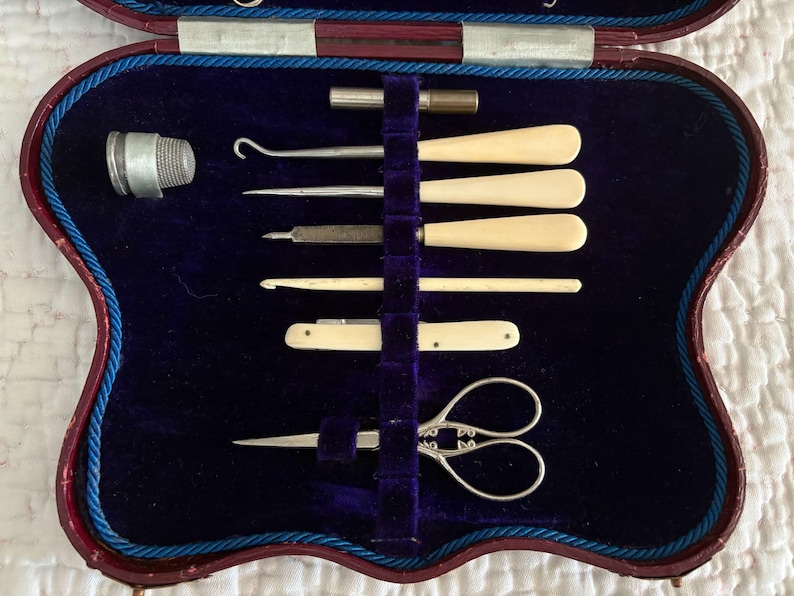May include: A vintage sewing kit in a burgundy case with a blue velvet interior. The kit includes a thimble, scissors, and various ivory-handled tools. The case is trimmed with blue cording.