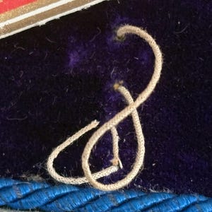 May include: Close-up of a section of a dark purple velvet surface with a cream-colored cord tied in a figure-eight shape. A blue braided trim is visible at the bottom. A portion of a red and gold label is in the upper left corner.