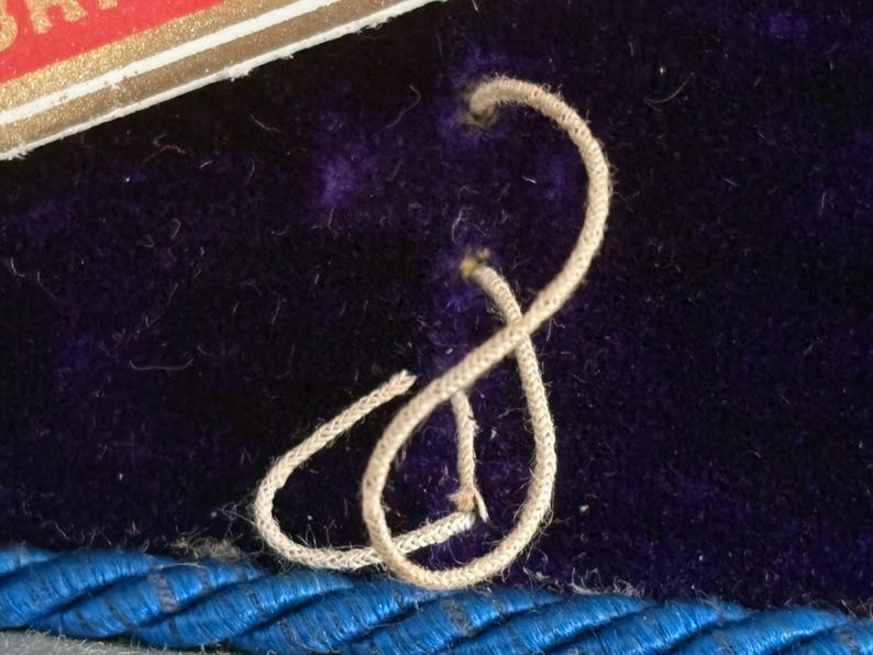 May include: Close-up of a section of a dark purple velvet surface with a cream-colored cord tied in a figure-eight shape. A blue braided trim is visible at the bottom. A portion of a red and gold label is in the upper left corner.