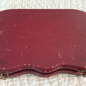 May include: A vintage burgundy-colored case with a textured surface. The case has a unique, scalloped shape and is closed with metal clasps. The case appears to be made of leather or a similar material, and shows signs of wear.