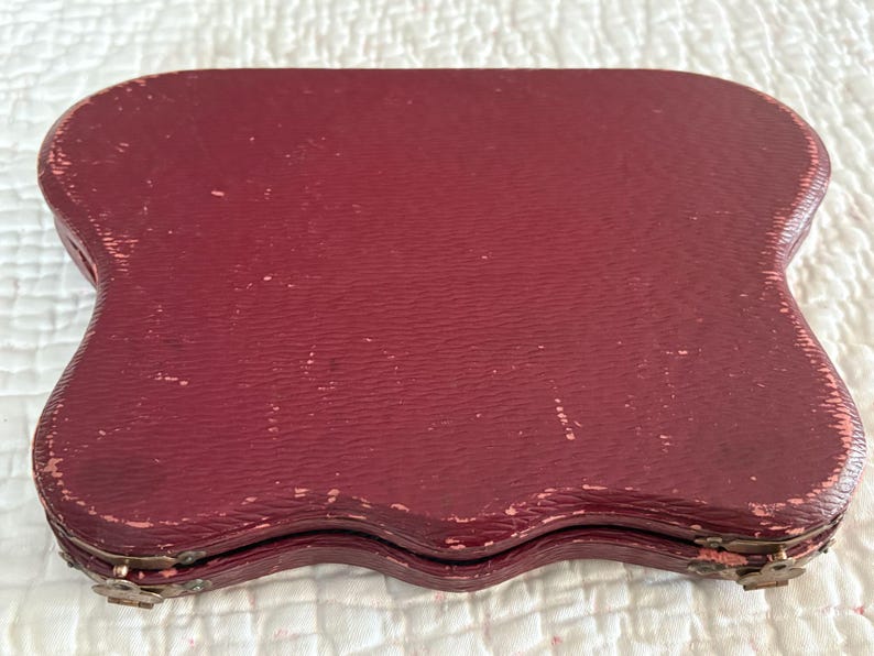 May include: A vintage burgundy-colored case with a textured surface. The case has a unique, scalloped shape and is closed with metal clasps. The case appears to be made of leather or a similar material, and shows signs of wear.