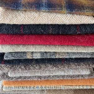 May include: A stack of various fabric swatches in shades of brown, red, black, and tan. The fabrics include plaid, herringbone, and solid textures. The swatches are stacked on top of each other, showcasing the different textures and colors.
