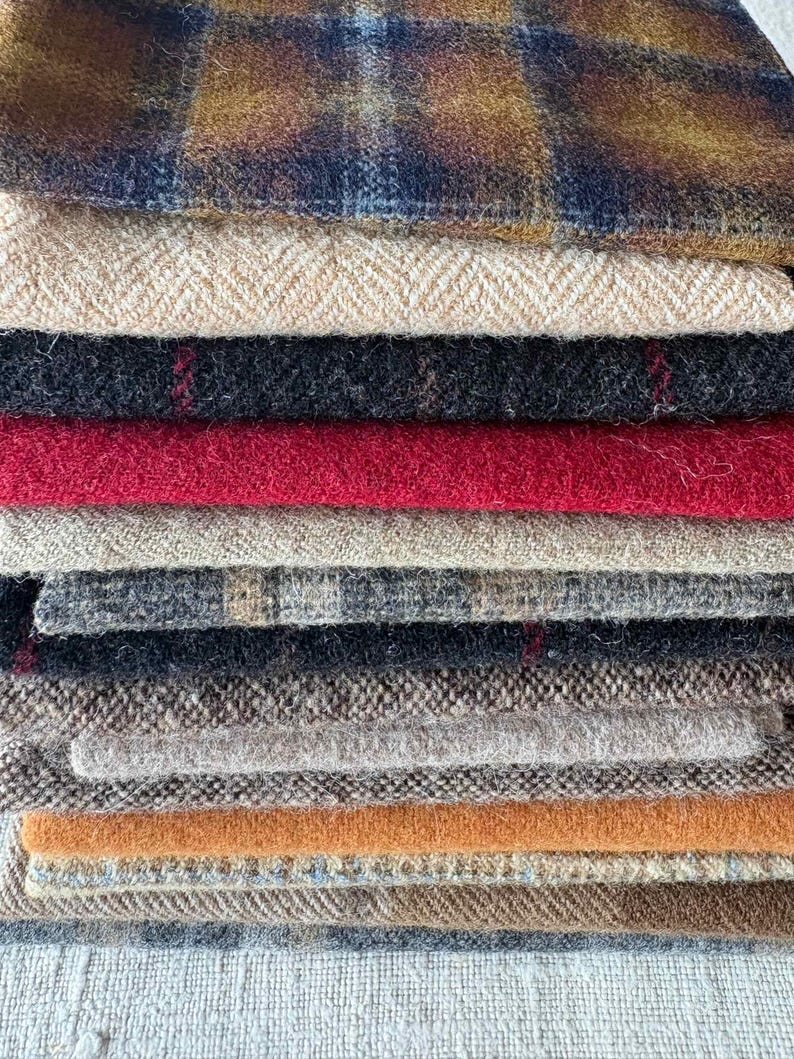 May include: A stack of various fabric swatches in shades of brown, red, black, and tan. The fabrics include plaid, herringbone, and solid textures. The swatches are stacked on top of each other, showcasing the different textures and colors.