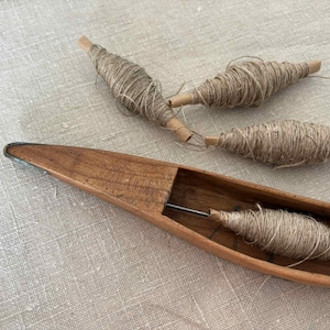 May include: A wooden boat-shaped weaving shuttle with a metal pin, and three spools of yarn. The spools are made of cardboard tubes wrapped with natural-colored yarn. The shuttle and spools are on a textured, neutral-colored surface.