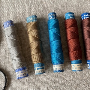 May include: Five spools of thread in various colours: grey, gold, teal, rust, and brown. The spools are labelled and sit next to a pair of vintage scissors. The thread is wound around the spools. The text on the spools is visible.