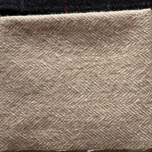 May include: Close-up of various fabric swatches. The primary swatch is a tan herringbone weave. Other swatches include a dark brown tweed, a black fabric with a red stripe, and a light gray fabric. The fabrics are likely for apparel or home decor.
