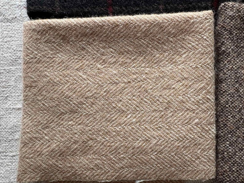 May include: Close-up of various fabric swatches. The primary swatch is a tan herringbone weave. Other swatches include a dark brown tweed, a black fabric with a red stripe, and a light gray fabric. The fabrics are likely for apparel or home decor.