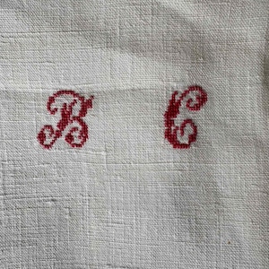 May include: Close-up of a cream-colored linen fabric with two embroidered red letters, "B" and "C". The letters are in a decorative, cursive style. The fabric has a subtle, textured weave.