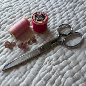 May include: A still-life image featuring sewing supplies. Two spools of thread, one pink and one red, sit next to a pair of silver Singer scissors. Dried flowers are scattered around the thread and scissors on a white quilted surface.