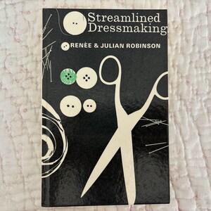 May include: A vintage book titled "Streamlined Dressmaking" by Renée & Julian Robinson. The cover is black with white illustrations of scissors, buttons, and pins. The buttons are white and green.
