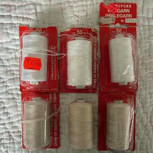 May include: Six spools of yarn in shades of white and beige are displayed. Each spool is packaged in a red cardboard backing with text. The packaging includes a price tag.