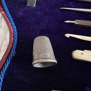 May include: Close-up of a vintage sewing kit with a silver thimble, needles, and other tools. The thimble is engraved with the word "DONALD". The tools are arranged on a dark blue velvet surface.