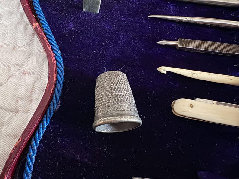 May include: Close-up of a vintage sewing kit with a silver thimble, needles, and other tools. The thimble is engraved with the word "DONALD". The tools are arranged on a dark blue velvet surface.