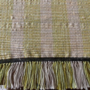 May include: Handwoven rectangular rug with a textured weave. The rug features a cream and yellow colour scheme with a black border and fringe. The fringe is a mix of yellow and cream. Suitable for home decor.