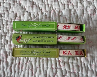 May include: Three green rectangular boxes containing white fabric tape with red embroidered initials. The boxes feature the text Une Grosse Garantie, INITIALES ET CHIFFRES POUR MARQUER LE LINGE, ARY, and Coton Grand Teint FABRICATION FRANÇAISE.