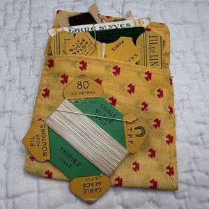 May include: A yellow fabric pouch with a small red geometric pattern containing several vintage cardboard spools of thread. One prominent green spool is labelled 80 20 METRES CABLE GLACE FIL POUR BOUTONS THIRIEZ LILLE. Other spools are partially visible.