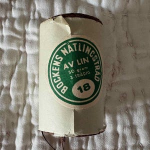 May include: A cylindrical spool of dark brown linen thread wrapped in a beige paper label. The label features a circular green emblem with the text BOCKENS NATLINGSTRAD, AV LIN, 50 gram, 3-TRADIG, and the number 18 printed in bold black ink.