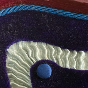May include: Close-up of a decorative object with a layered design. The top layer is a dark purple velvet, edged with a blue twisted cord. Below is a light blue, pleated fabric, and a blue button.