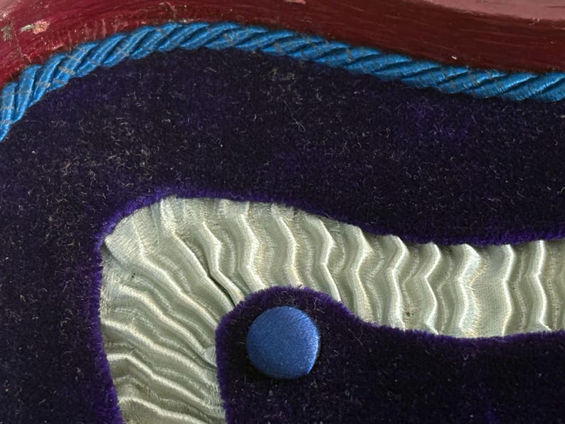 May include: Close-up of a decorative object with a layered design. The top layer is a dark purple velvet, edged with a blue twisted cord. Below is a light blue, pleated fabric, and a blue button.