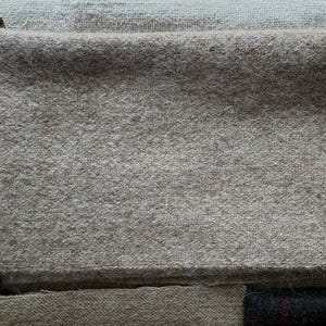 May include: A folded square of light gray wool fabric, possibly for sewing or crafting. The fabric has a soft, textured appearance. Other fabric swatches and buttons are visible in the background.