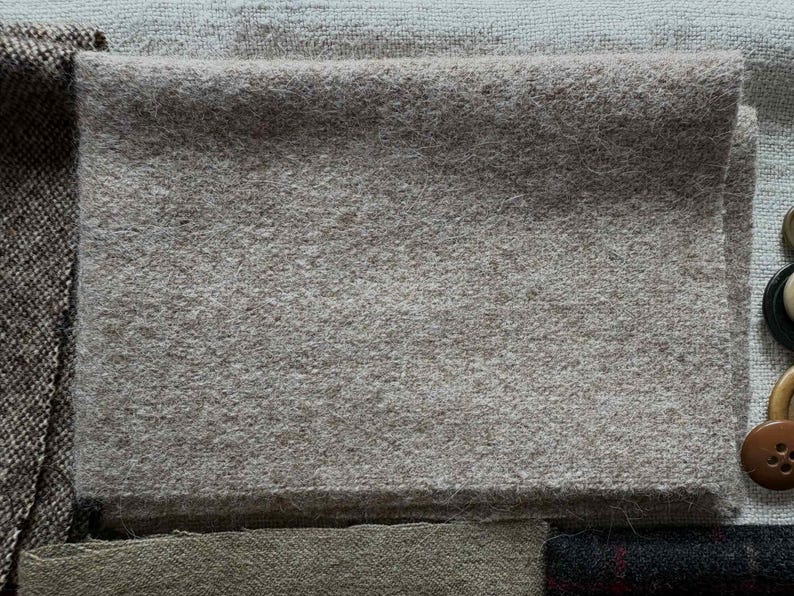 May include: A folded square of light gray wool fabric, possibly for sewing or crafting. The fabric has a soft, textured appearance. Other fabric swatches and buttons are visible in the background.