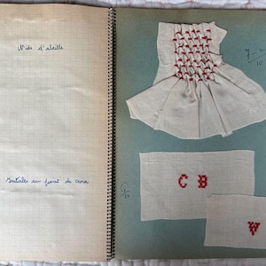 May include: A notebook with a spiral binding, displaying fabric samples with red cross-stitch embroidery. One sample is a small garment, and others show initials. Handwritten text in blue ink is also visible.