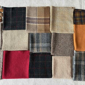 May include: A collection of fabric swatches in various colors and patterns, including plaid, solid, and textured designs. The color palette includes shades of brown, gray, red, and orange. A pile of assorted buttons is in the upper left corner.