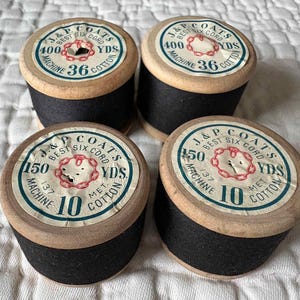 May include: Four vintage spools of black thread. Each spool has a wooden base and a paper label with text including "J&P Coats Best Six Cord" and yardage measurements. The labels have a red and white design.