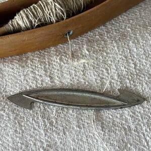 May include: A wooden weaving shuttle containing a spool of beige thread, with a thin thread extending from the shuttle to a separate, elongated, pointed metal tool with a central groove and hooked ends, resting on a textured fabric surface.