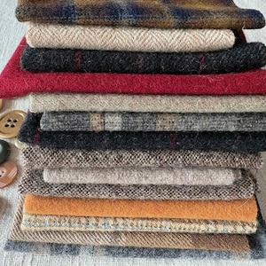 May include: A stack of various fabric swatches in shades of brown, red, black, and beige. The fabrics have different textures and patterns, including herringbone and plaid. Several brown and black buttons are scattered to the left of the fabric stack.