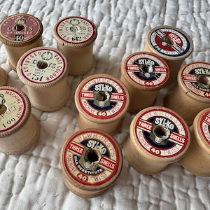 May include: A collection of twelve vintage wooden sewing thread spools with circular paper labels on top. The labels feature various brand names including Coats and Dewhurst's Sylko, along with colour names, numbers, and machine twist specifications.