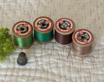 May include: Four vintage spools of thread in green, dark green, burgundy, and tan, with a silver thimble. The spools have red and white labels with the text "SYLKO" and "DEWHURST'S". A sewing-themed image.