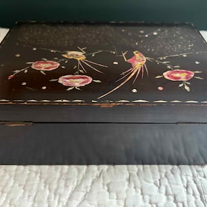 May include: A black wooden box with a hinged lid. The lid is decorated with pink flowers, birds, and white dots. The box has a rectangular shape and is likely used for storage. The box is sitting on a white, textured surface.